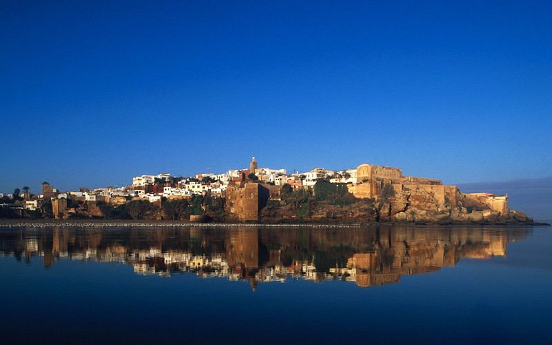 Cette magnifique photographie met en valeur la Kasbah des Oudayas, l'un des sites historiques les plus emblématiques de Rabat. Baignée par la douce lumière du crépuscule, la forteresse aux remparts ocre se reflète parfaitement dans les eaux calmes du fleuve Bouregreg, créant une scène d'une rare sérénité.